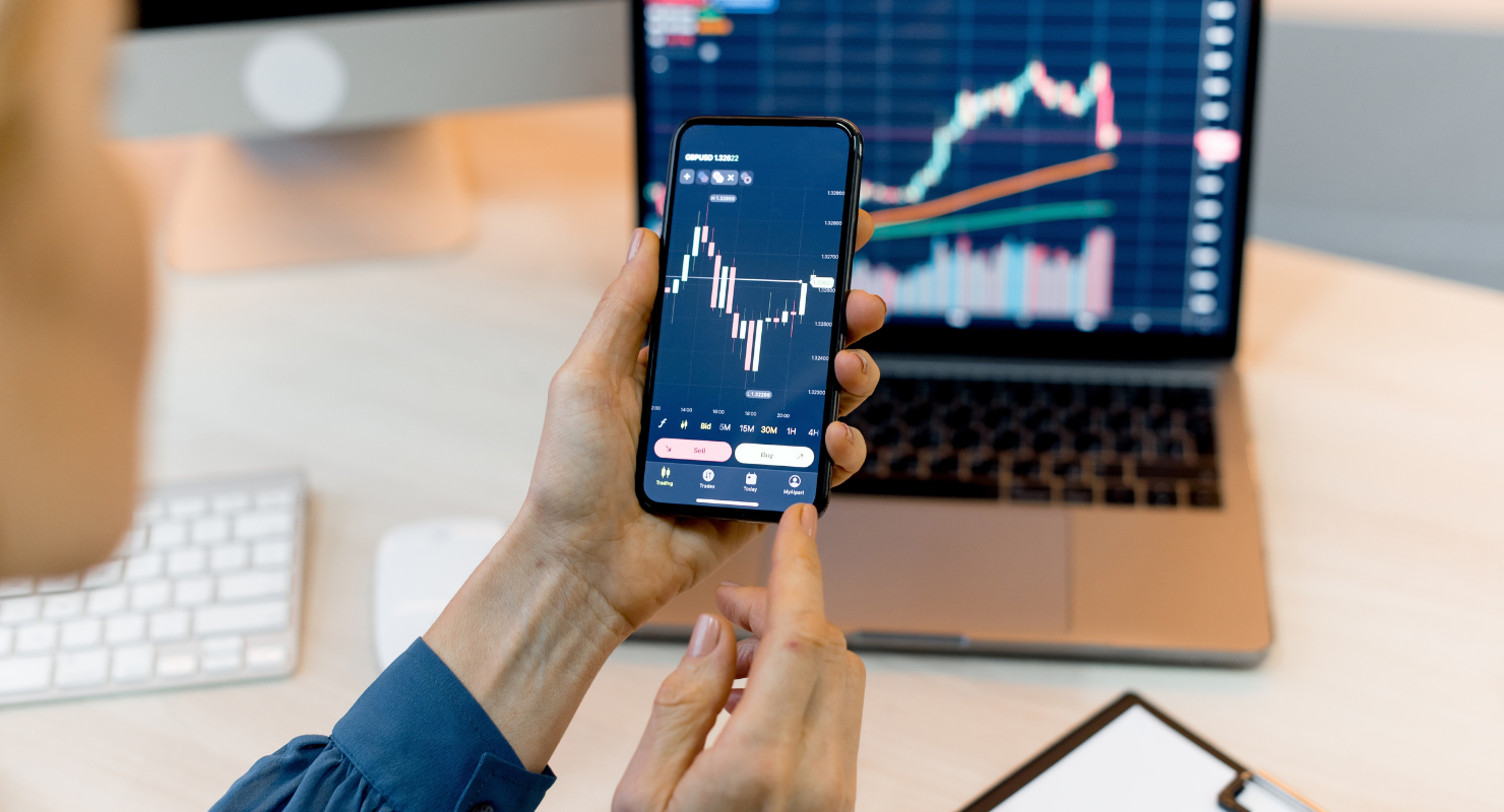 Hands holding a phone with stock charts, a computer with financial data in the background.
